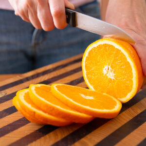 Woman standing near table and cutting orange on slices with knife on wooden board. Ripe citrus fruit. Cropped side view. Studio shot. Nutrition and vegetarian concept