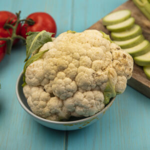 top view of fresh cauliflower on a bowl with chopped zucchinis on a wooden kitchen board with celery with tomatoes isolated on a blue wooden background